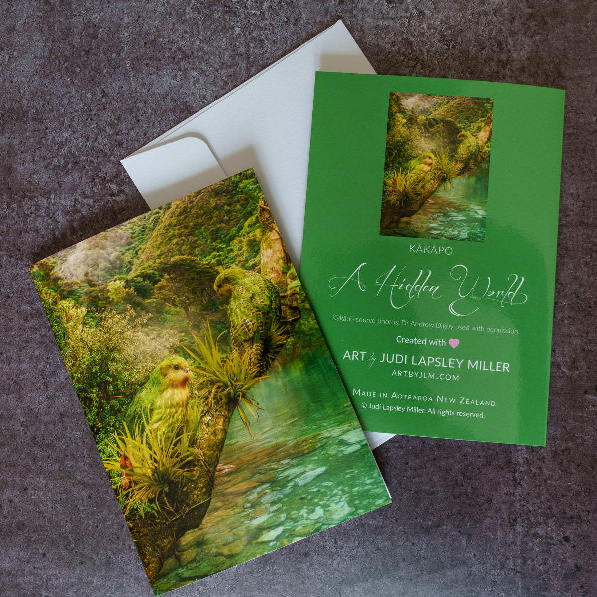 Greeting cards and envelope arranged on a grey surface. The card features two kakapo in a forest scene over water.
