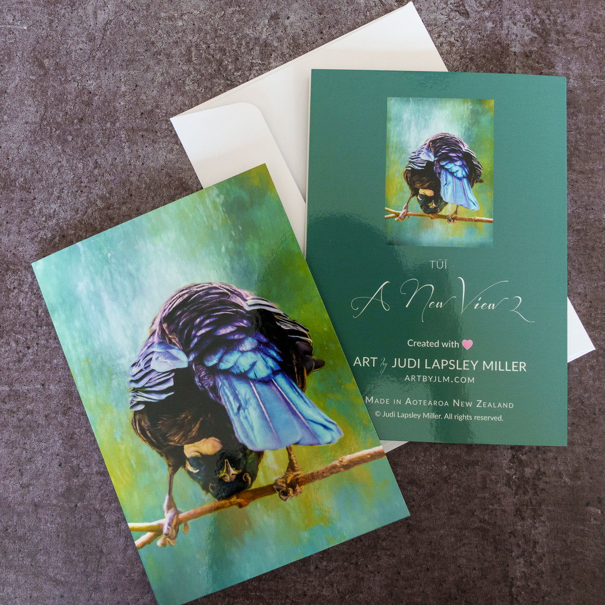 Two art prints and a white envelope arranged on a textured gray surface. The prints feature a painted blue and black bird perched on a branch against green and yellow tones, with one print showing the full artwork and the other showing a smaller preview with printed text.