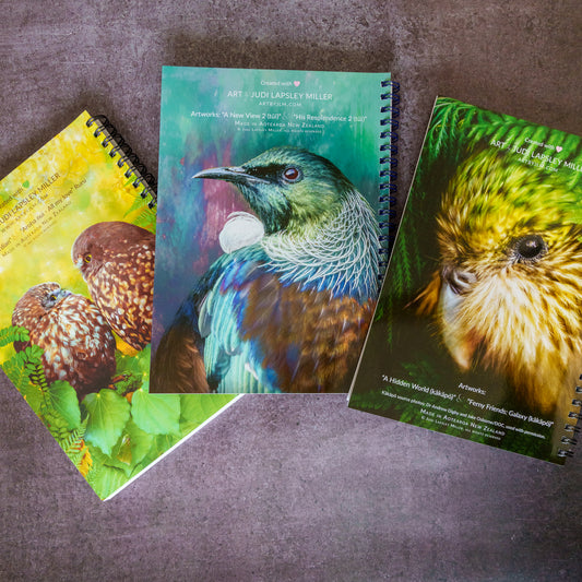 Three spiral-bound art notebooks featuring colorful bird illustrations are arranged on a textured gray surface. The center notebook shows a teal and blue bird with the text 'ART by JUDI LAPSLEY MILLER' .