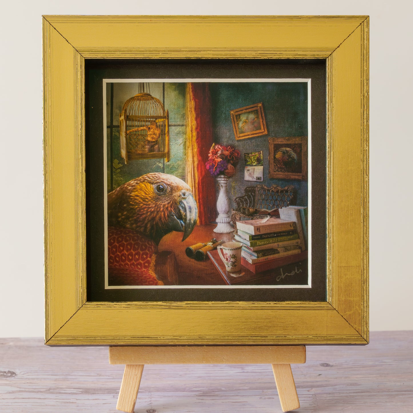 A framed art print featuring a detailed still life painting of a parrot in a room with books, flowers, and a birdcage, displayed on a small wooden easel against a light wall.