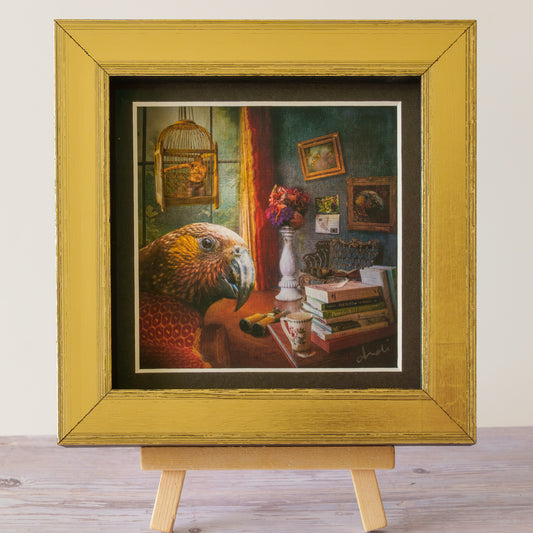 A framed art print featuring a detailed still life painting of a parrot in a room with books, flowers, and a birdcage, displayed on a small wooden easel against a light wall.