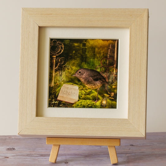 A framed art print featuring a small brown bird perched on mossy ground with a book and surreal architectural elements, displayed in a light wood frame on a small wooden easel against a neutral background.