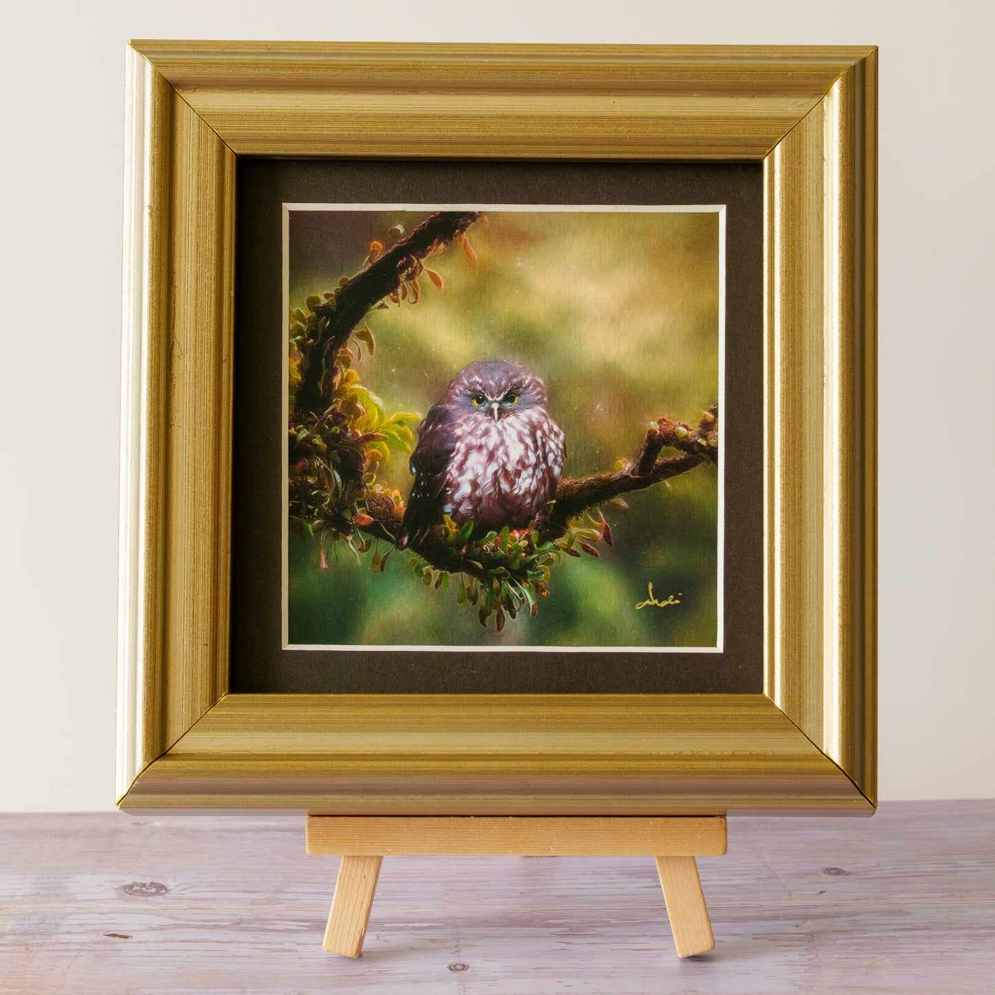 A framed owl painting displayed on a small wooden easel. The artwork features a small owl perched on a branch with a soft, blurred green and yellow background.
