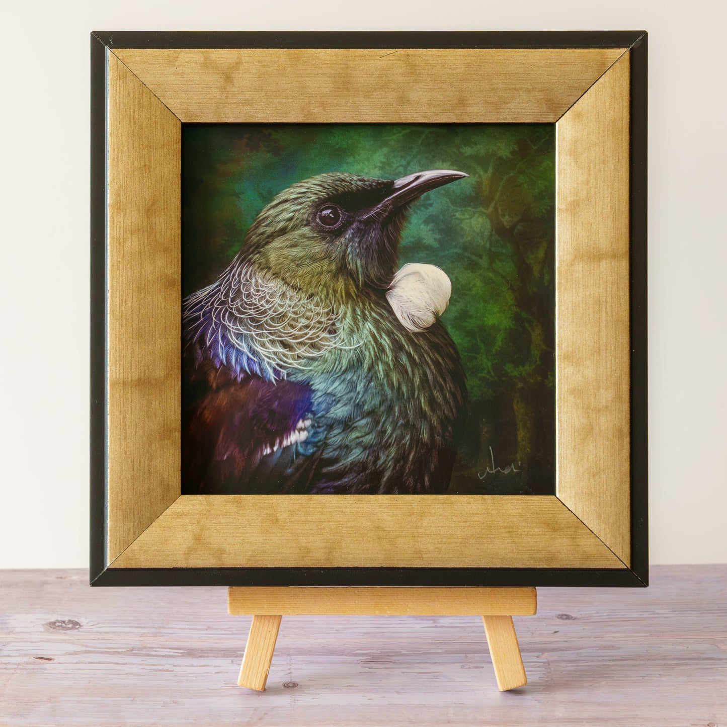 A framed artwork of a bird with iridescent green, blue, and purple feathers, displayed on a small wooden easel against a light wall and wood surface.