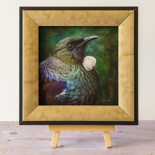 A framed artwork of a bird with iridescent green, blue, and purple feathers, displayed on a small wooden easel against a light wall and wood surface.