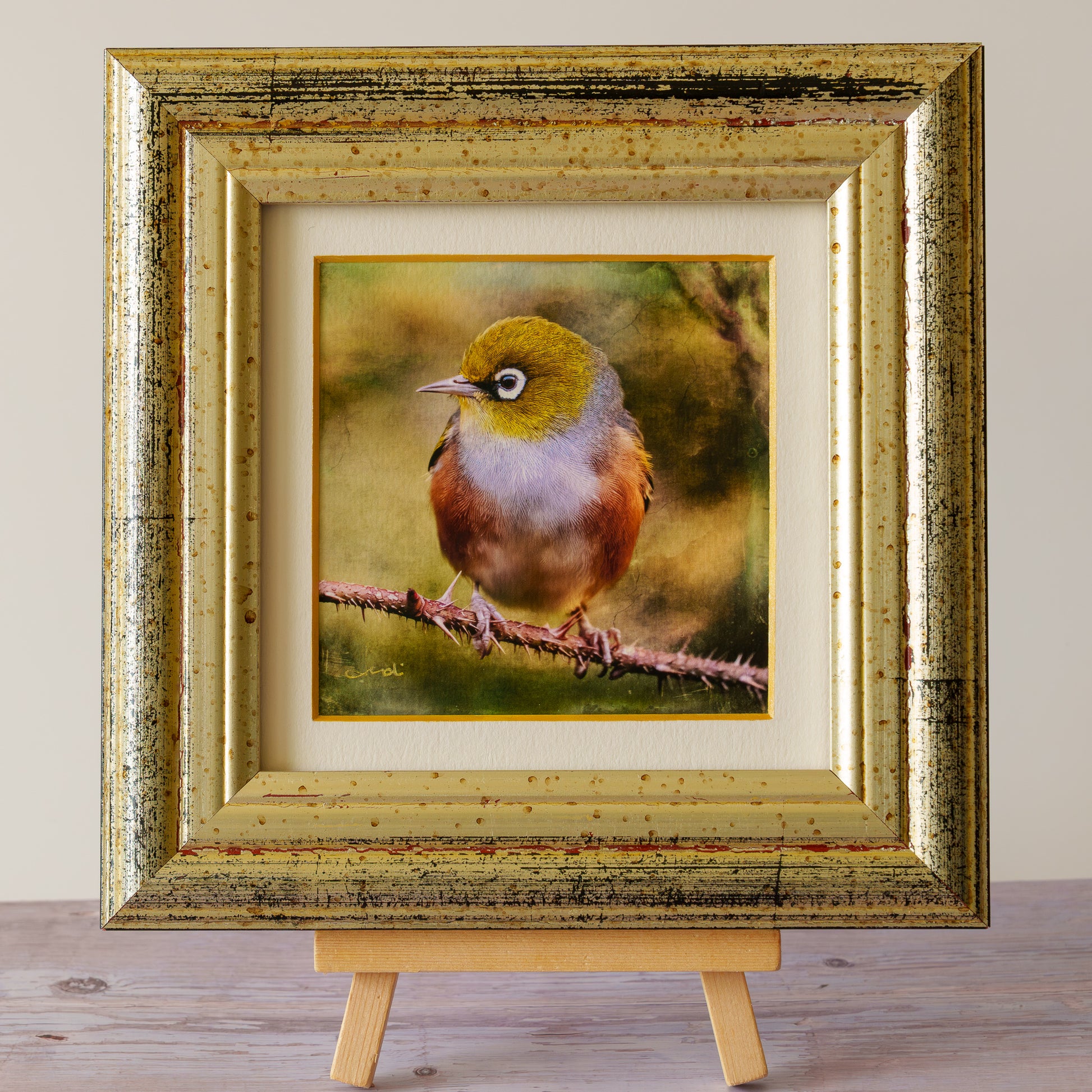 A framed bird artwork displayed on a small wooden easel. The print shows a small brown and olive bird perched on a branch, set within a cream mat and a distressed gold frame, against a neutral indoor background.