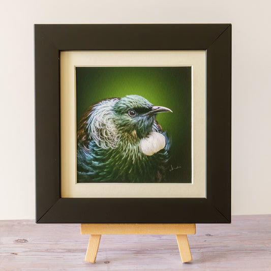 A framed bird painting displayed on a small wooden easel against a light beige wall and wood tabletop. The artwork shows a green and blue bird with a white chest, set within cream matting and a black frame.
