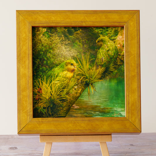 A square framed artwork on a wooden easel, showing two yellow-green parrots in a lush green forest beside a turquoise body of water. The frame is a warm golden wood tone against a light neutral wall.