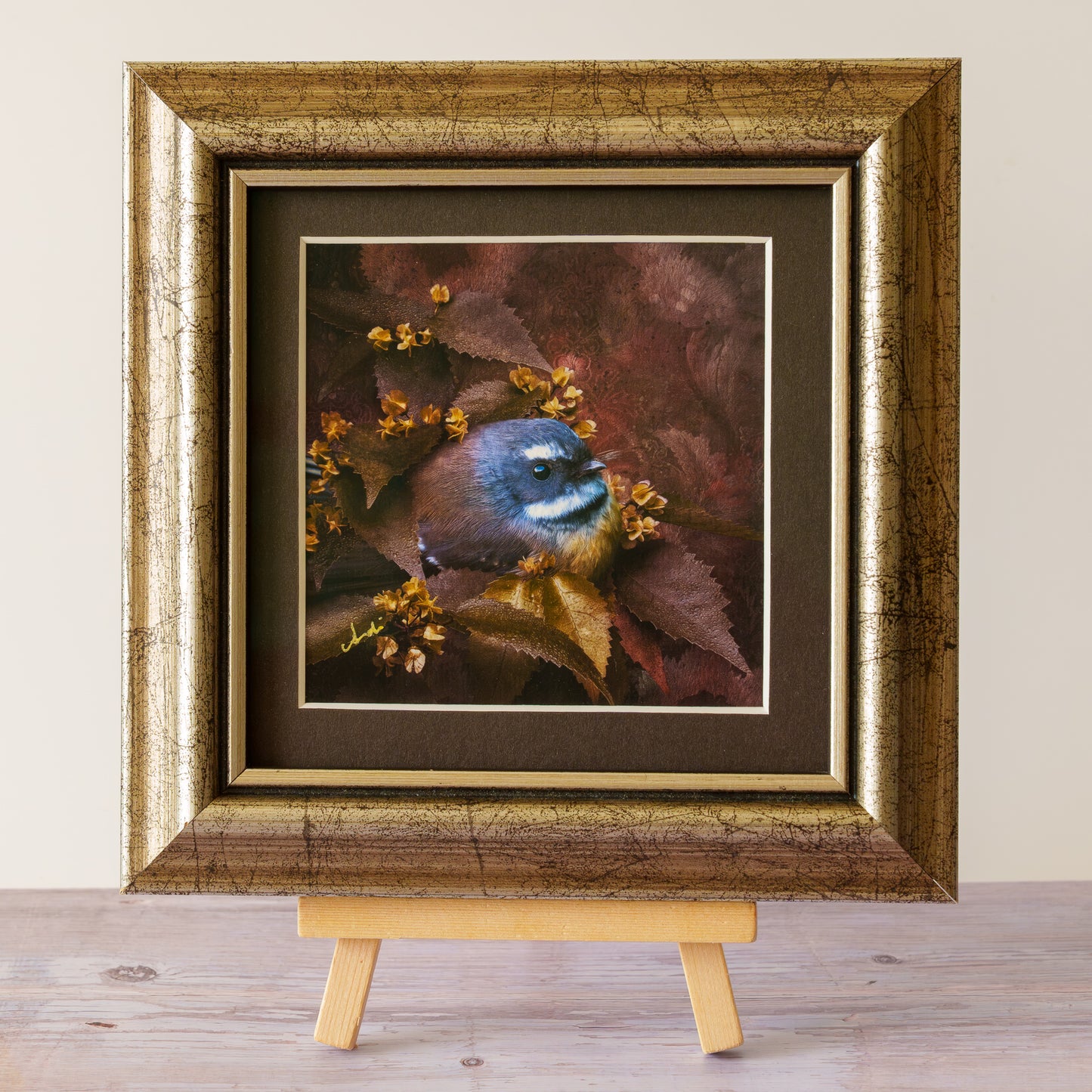 A framed art print of a small bird perched among brown and gold leaves, displayed on a small wooden easel against a neutral beige wall and light wood floor.