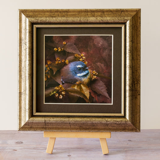 A framed art print of a small bird perched among brown and gold leaves, displayed on a small wooden easel against a neutral beige wall and light wood floor.