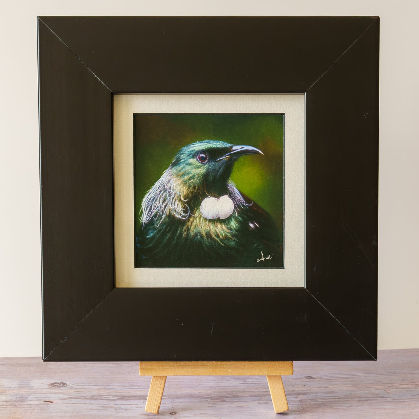 A framed bird painting displayed on a small wooden easel. The artwork shows a green and black bird with a white chest patch against a dark green background, set within a cream mat and a thick black frame.