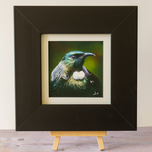 A framed bird painting displayed on a small wooden easel. The artwork shows a green and black bird with a white chest patch against a dark green background, set within a cream mat and a thick black frame.