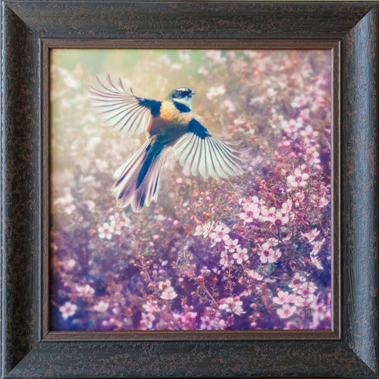 Framed square wall art featuring a small bird in flight over pink and purple flowering plants, set in a dark textured wooden frame.