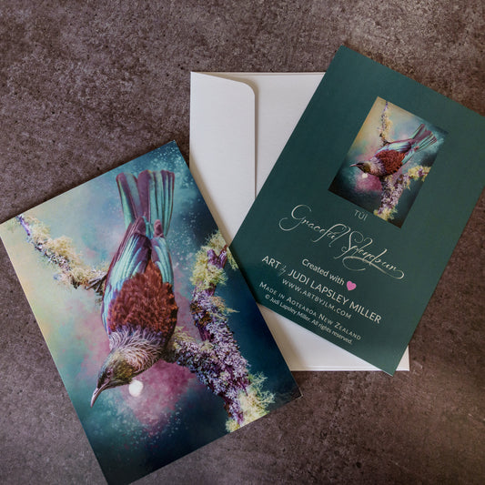A custom printed greeting card and matching envelope laid on a textured gray surface. The card features a colorful painted tui bird perched on flowering branches, with the back showing the text 'TUI Graceful Splendour' and creator details.