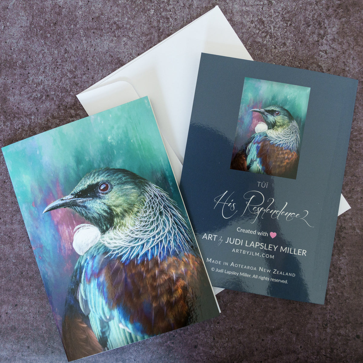Two art cards featuring a colorful painted bird portrait, arranged on a textured purple surface with a white envelope behind them. One card shows the front artwork, and the other shows the back with the title 'His Resplendence' and artist credit for Judi Lapsley Miller.