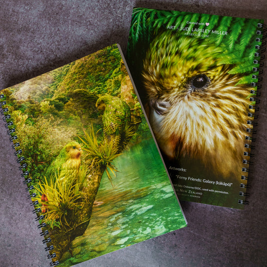 Two spiral-bound notebooks featuring detailed nature artwork of green parrots in a forest scene, displayed on a textured gray surface.