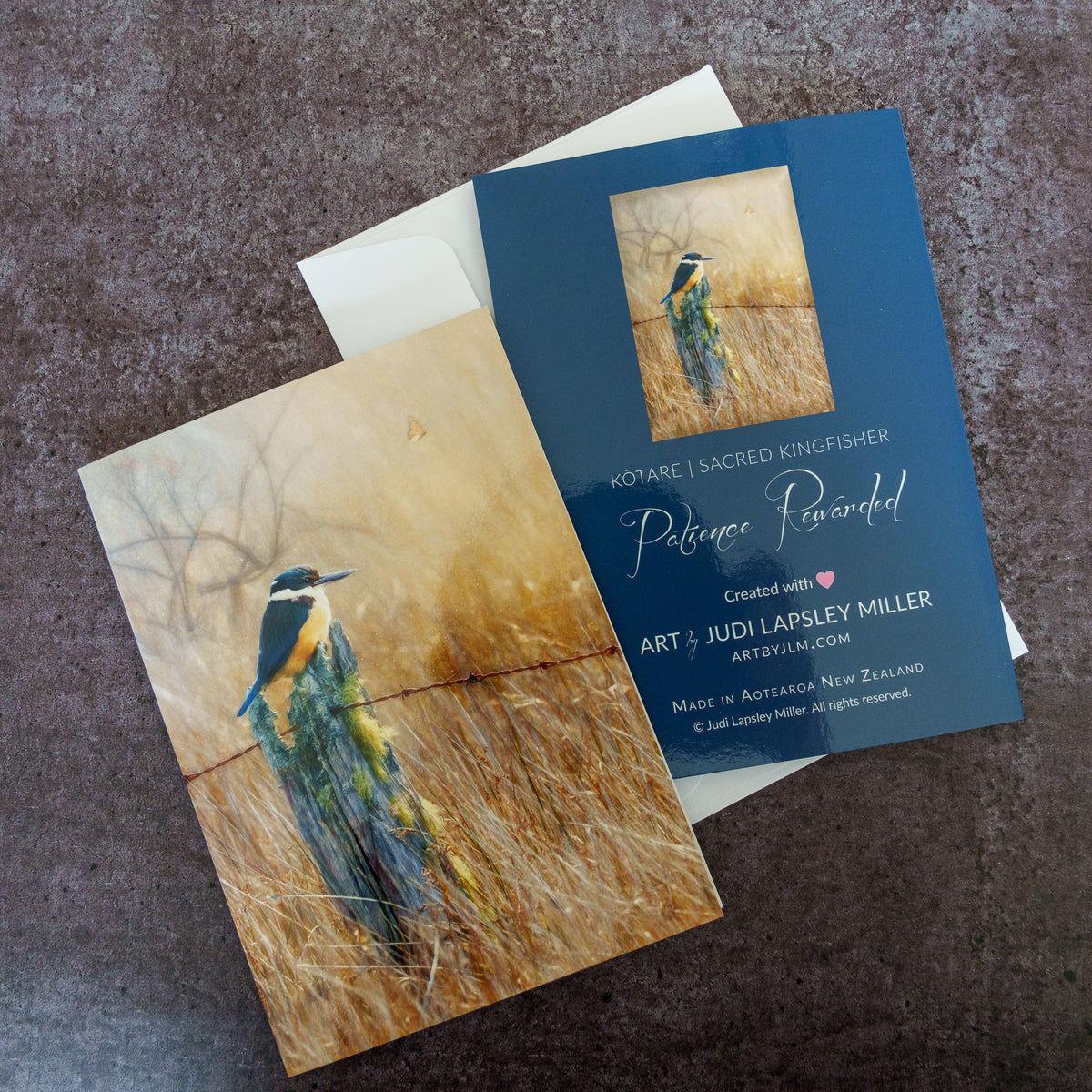Two art greeting cards featuring a painted kingfisher in a grassy field, arranged on a textured dark surface. One print is a warm beige-toned painting, and the other is dark blue with white text reading 'KOTARE | SACRED KINGFISHER' and 'Patience Rewarded' plus artist credit 'ART BY JUDI LAPSLEY MILLER'.