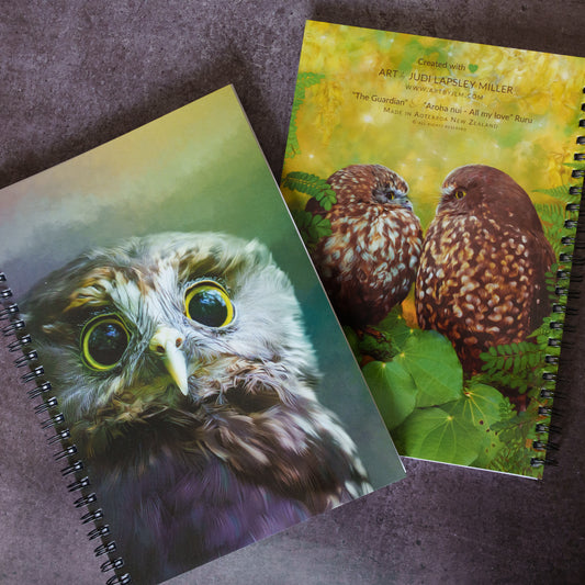 Two spiral-bound notebooks featuring detailed owl artwork, displayed on a textured dark gray surface. One notebook shows a close-up of an owl with large yellow eyes, and the other shows two brown owls under a yellow-green sky.