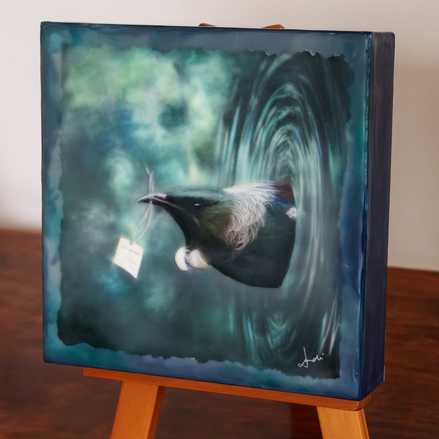 Wax finished mixed media artwork of a tui coming out of a vortex carrying a specimen label in his beak. Angled view.