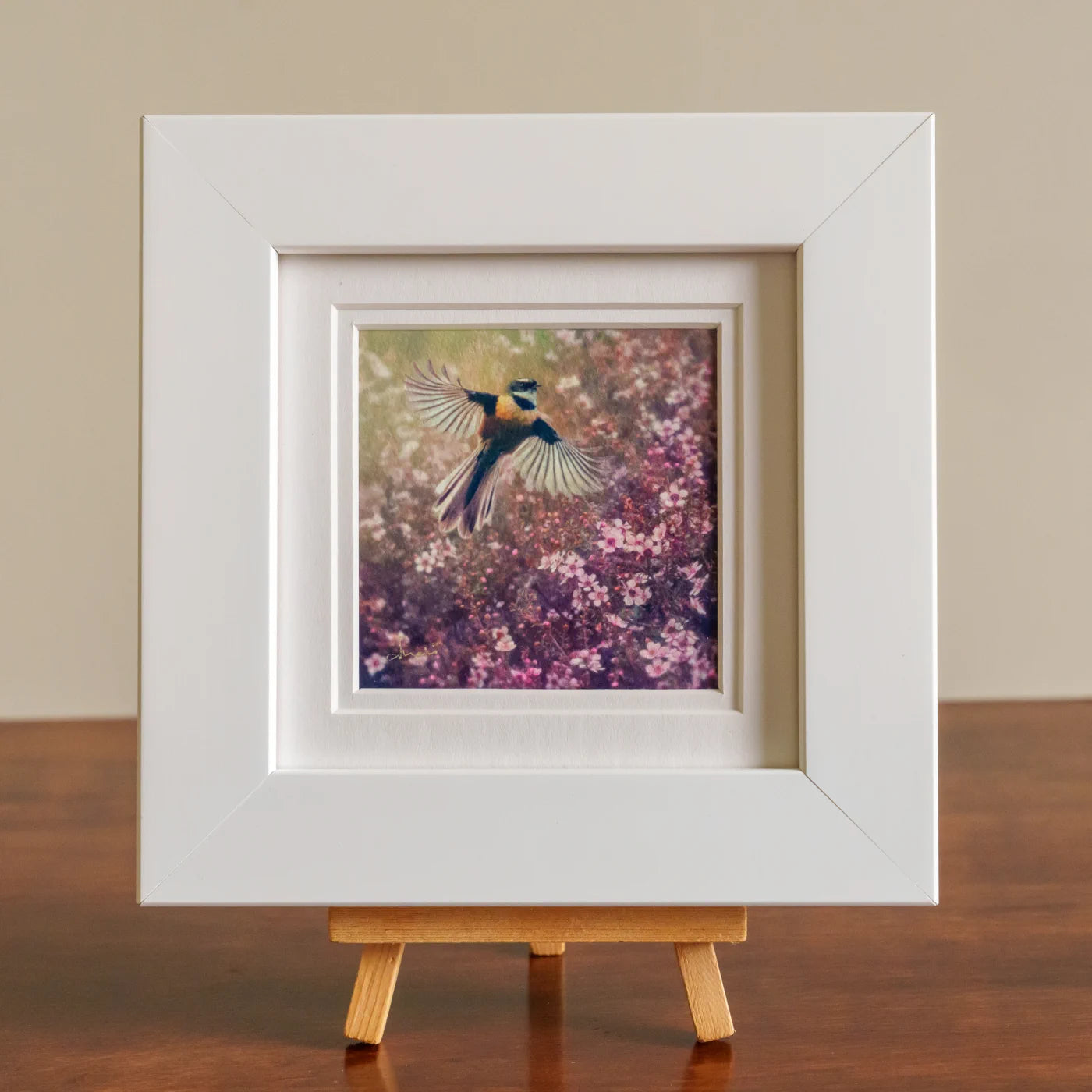 Framed artwork of a bird in flight over flowers on a wooden stand.