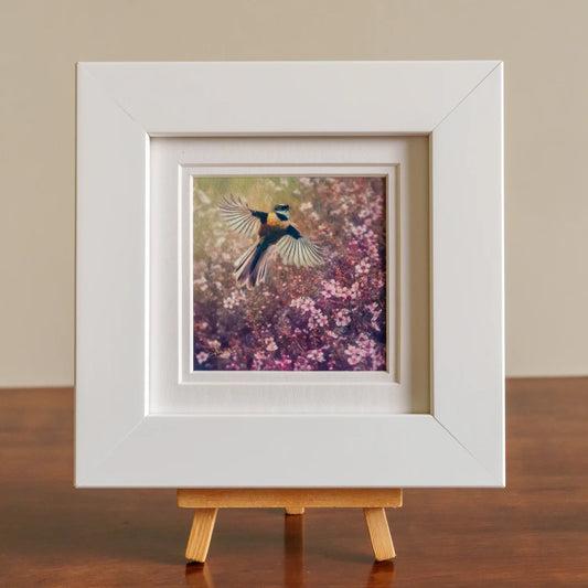 Framed artwork of a bird in flight over flowers on a wooden stand.