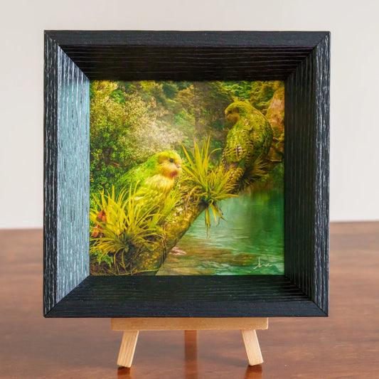 Framed artwork of two parrots in a lush green forest setting on a wooden stand.