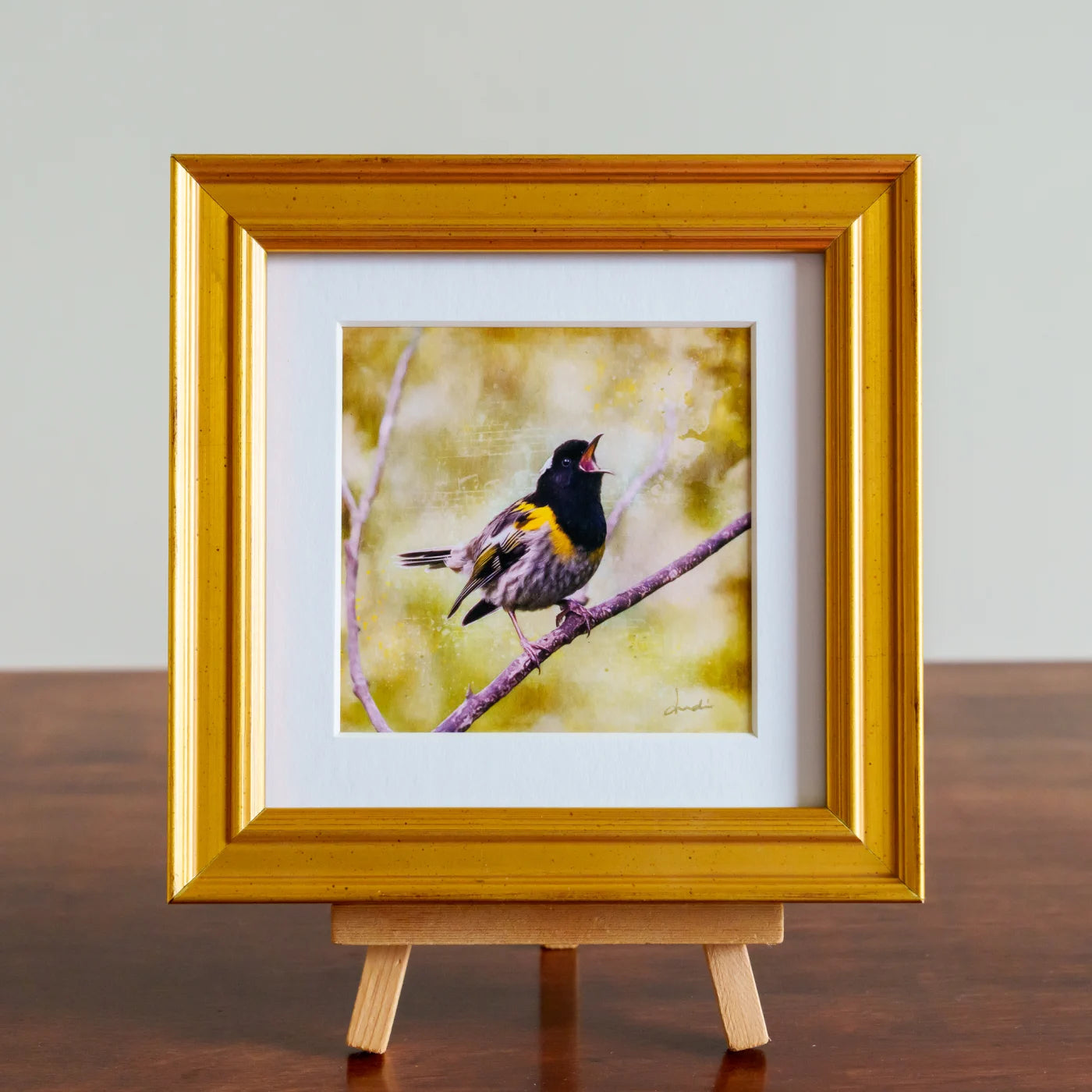 Framed artwork of a bird on a branch with a gold frame on a wooden stand.