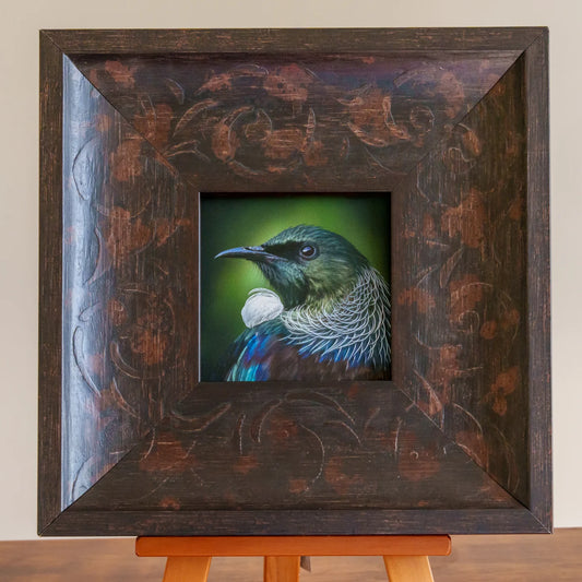 Framed artwork of a colorful bird on an easel against a plain wall.