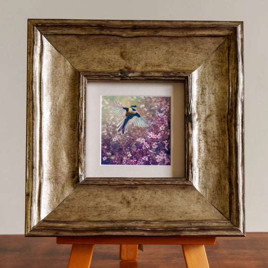 Framed artwork of a bird on a floral background in a metallic frame on an easel.