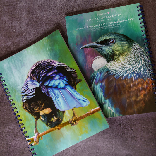 Two spiral-bound art notebooks featuring colorful bird illustrations, placed on a textured brown surface. One notebook shows a blue and purple bird upside down on a branch, and the other shows a green and brown bird with a white neck patch.