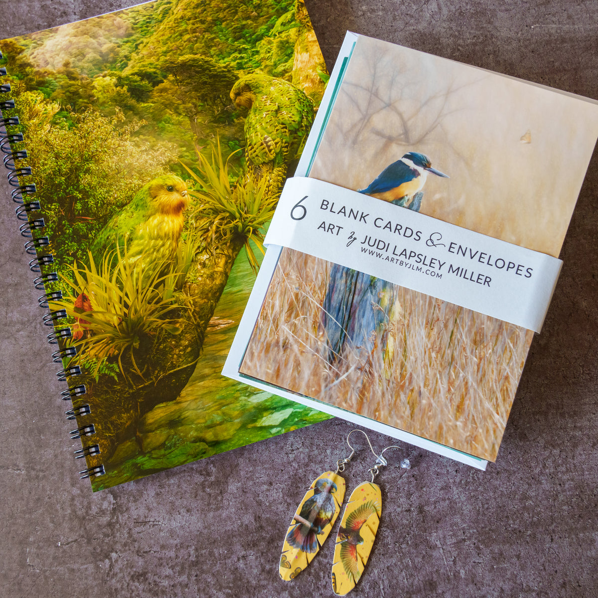 A spiral-bound art notebook and a pack of blank cards and envelopes from Art & Judi Lapsley Miller, styled on a textured brown surface with yellow kaka parrot earrings in front.
