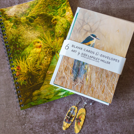 A spiral-bound art notebook and a pack of blank cards and envelopes from Art & Judi Lapsley Miller, styled on a textured brown surface with yellow kaka parrot earrings in front.
