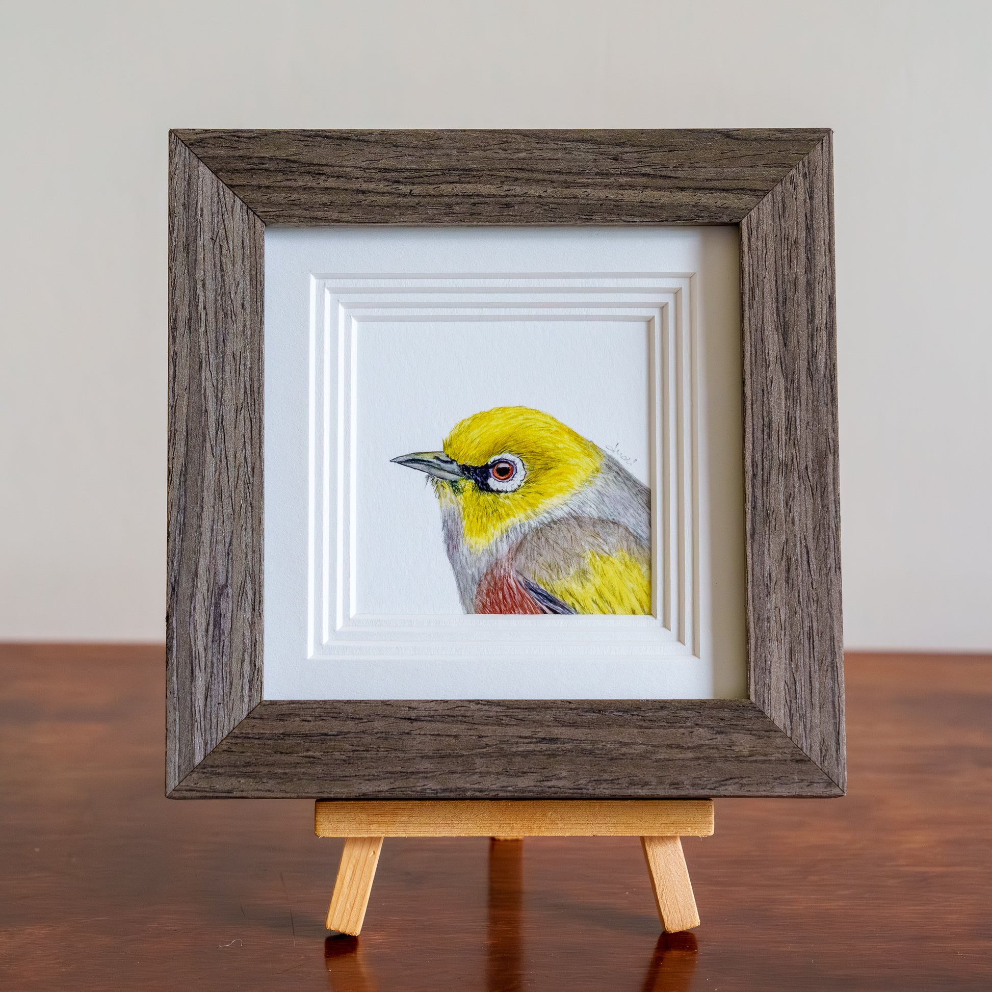 Framed painting of a colorful bird on a wooden easel against a neutral background