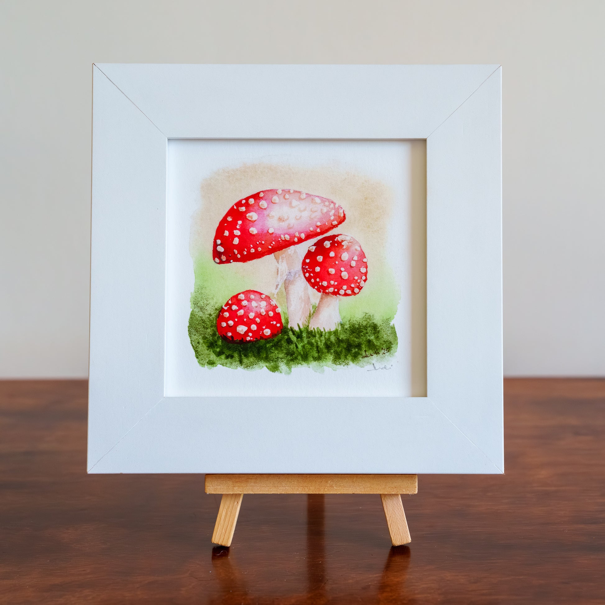 Framed artwork of red mushrooms on a wooden stand against a neutral background
