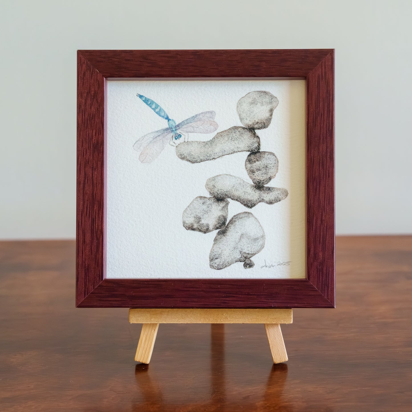 Framed artwork of stones and a dragonfly on a wooden stand