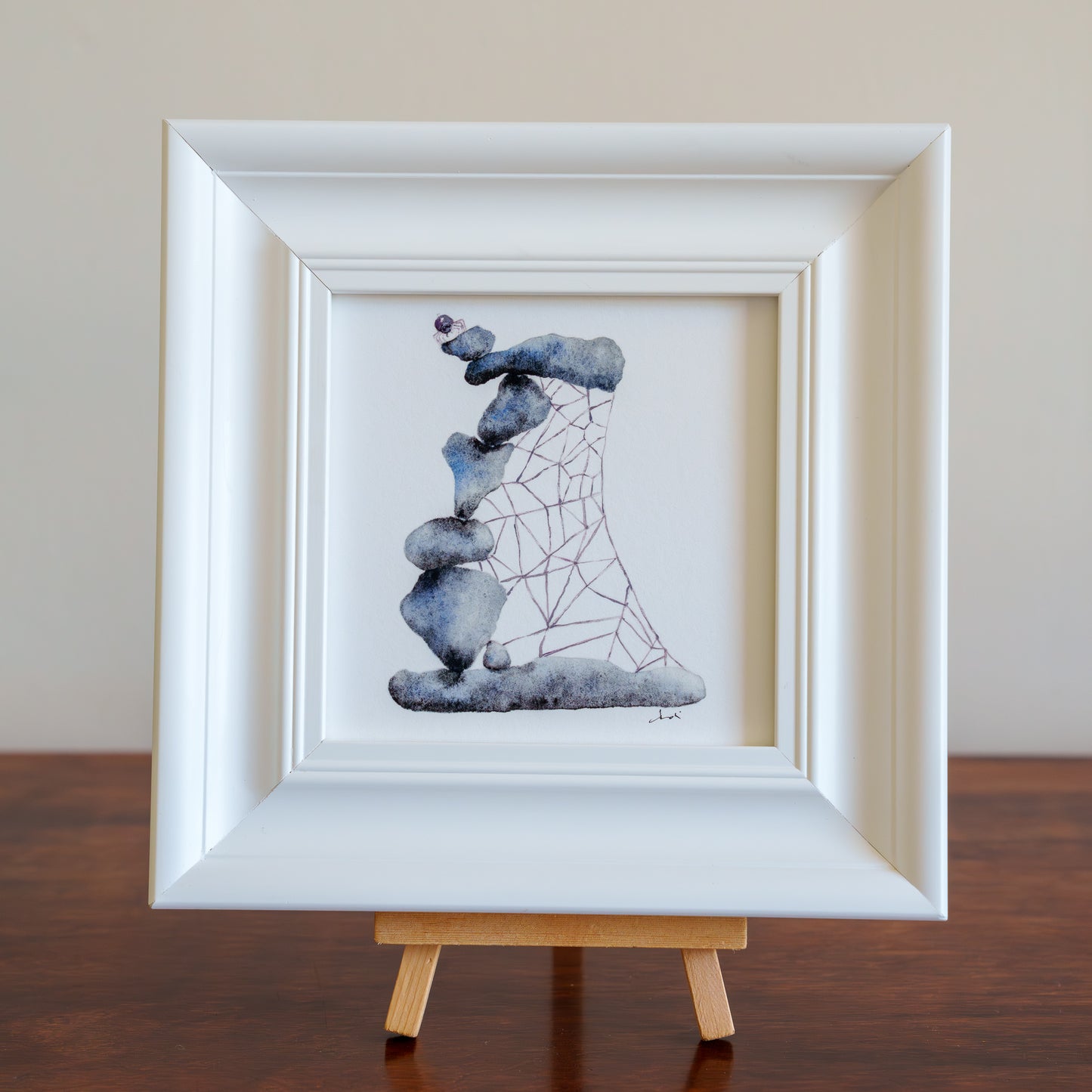 Framed artwork of stacked stones with a spiderweb on a wooden stand against a plain background