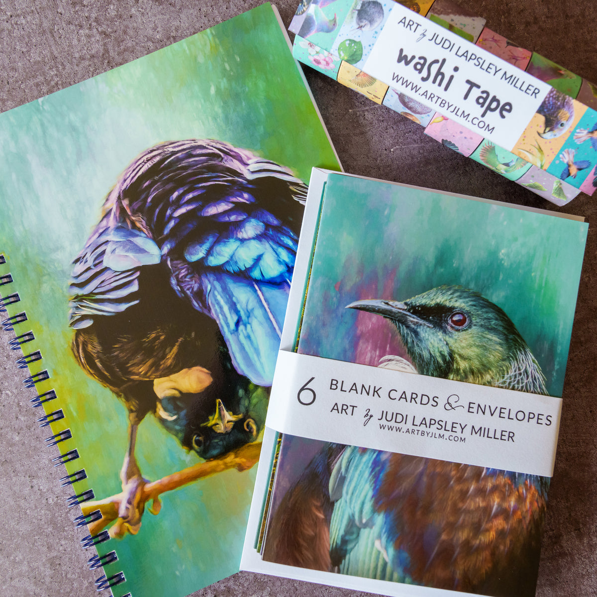 A styled flat-lay of art supplies on a textured brown surface, featuring a spiral-bound notebook with a colorful bird illustration, a stack of blank cards and envelopes wrapped in a paper band reading '6 Blank Cards & Envelopes Art by Judi Lapsley Miller', and a roll of washi tape labeled 'Art by Judi Lapsley Miller washi Tape'.