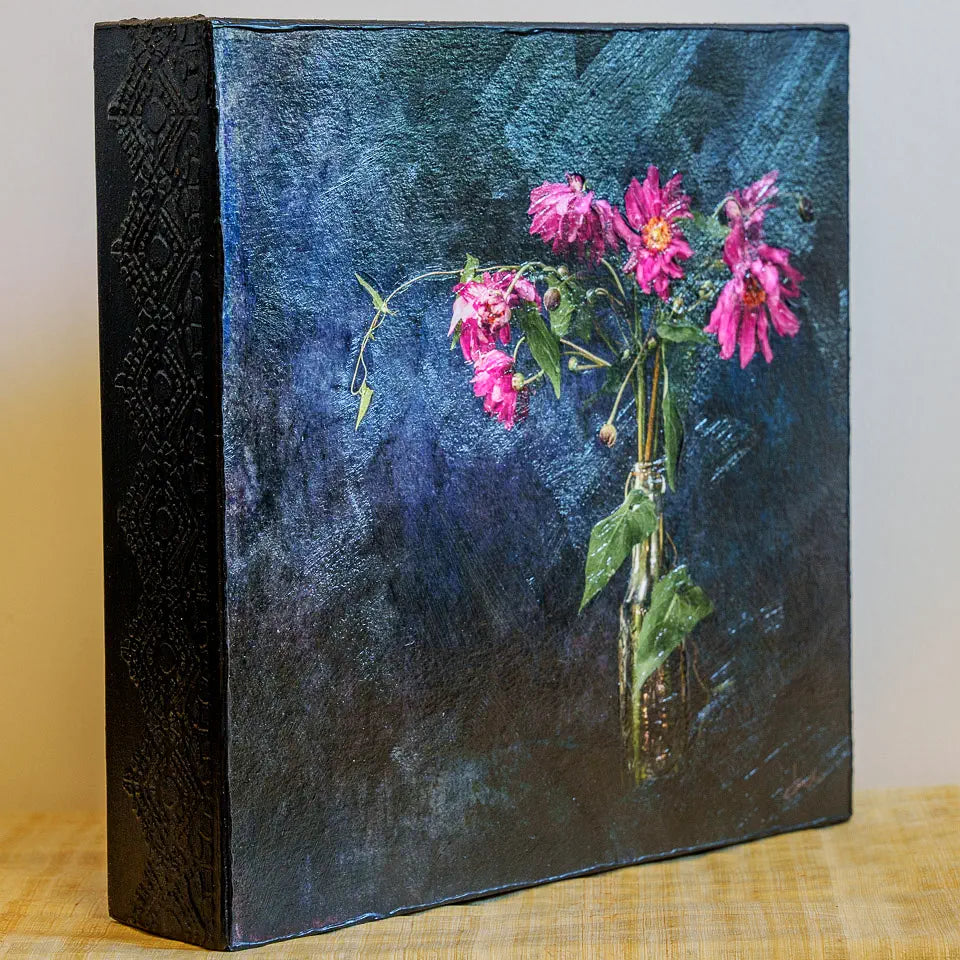 Photo of a mounted artwork of flowers in a bottle with a dark textured background. This is an angled view showing the lacey embossed sides of the cradle board and the brushwork texture of the impasto finish