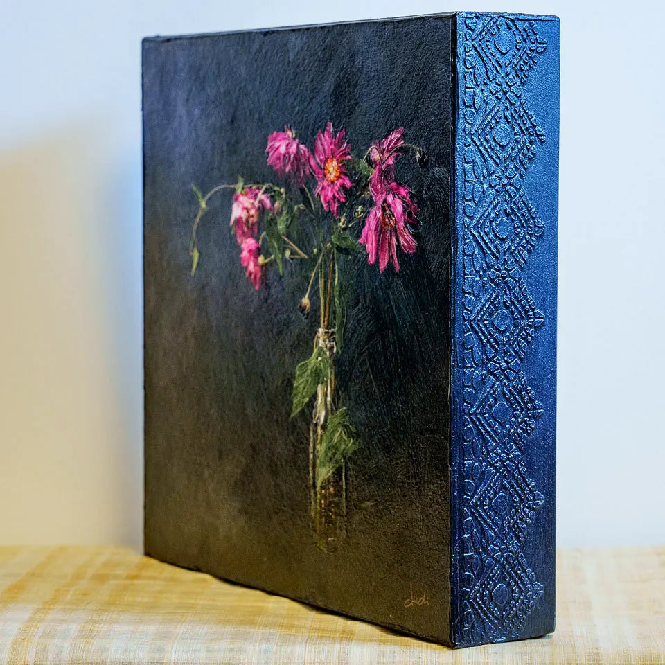 Photo of a mounted artwork of flowers in a bottle with a dark textured background. This is an angled view showing the lacey embossed sides of the cradle board and the brushwork texture of the impasto finish