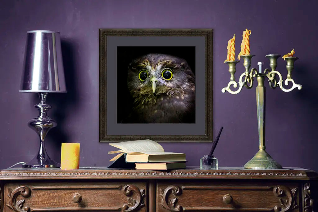 Side table with lamps, books, and candles and a framed ruru owl artwork against a purple wall