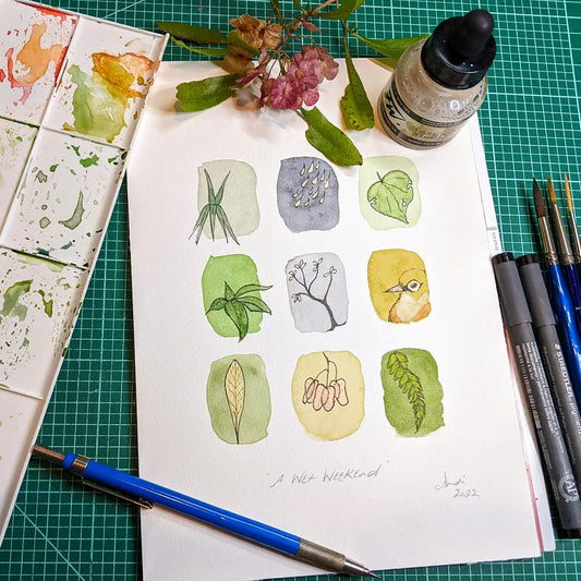 Photo of a watercolour painting of leaves and a bird, surrounded by art supplies and a sprig of akeake