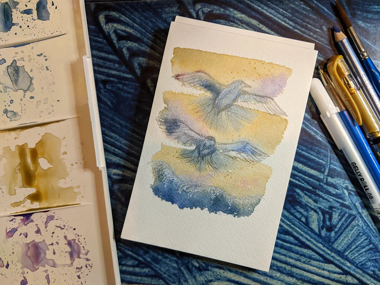 Photo of a watercolour painting of two gulls flying above a breaking wave. There are art supplies surrounding the painting.