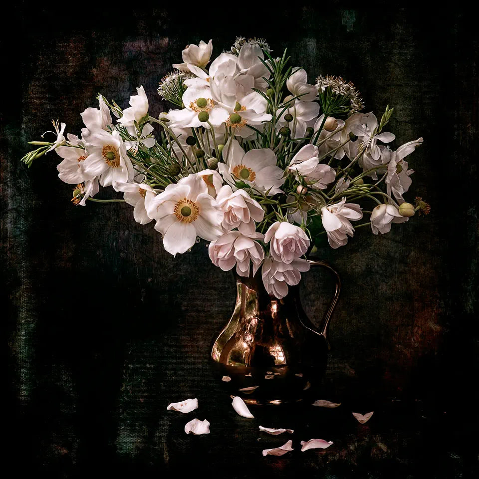 A still life photograph of white flowers and herbs in a jug with fallen petals